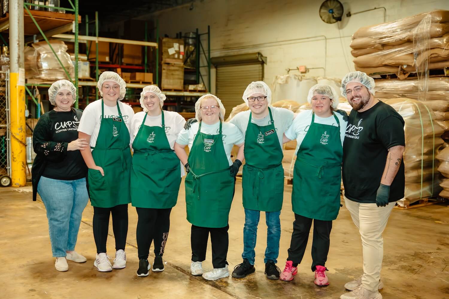 Seven CUP team members in green aprons posed together at the roastery