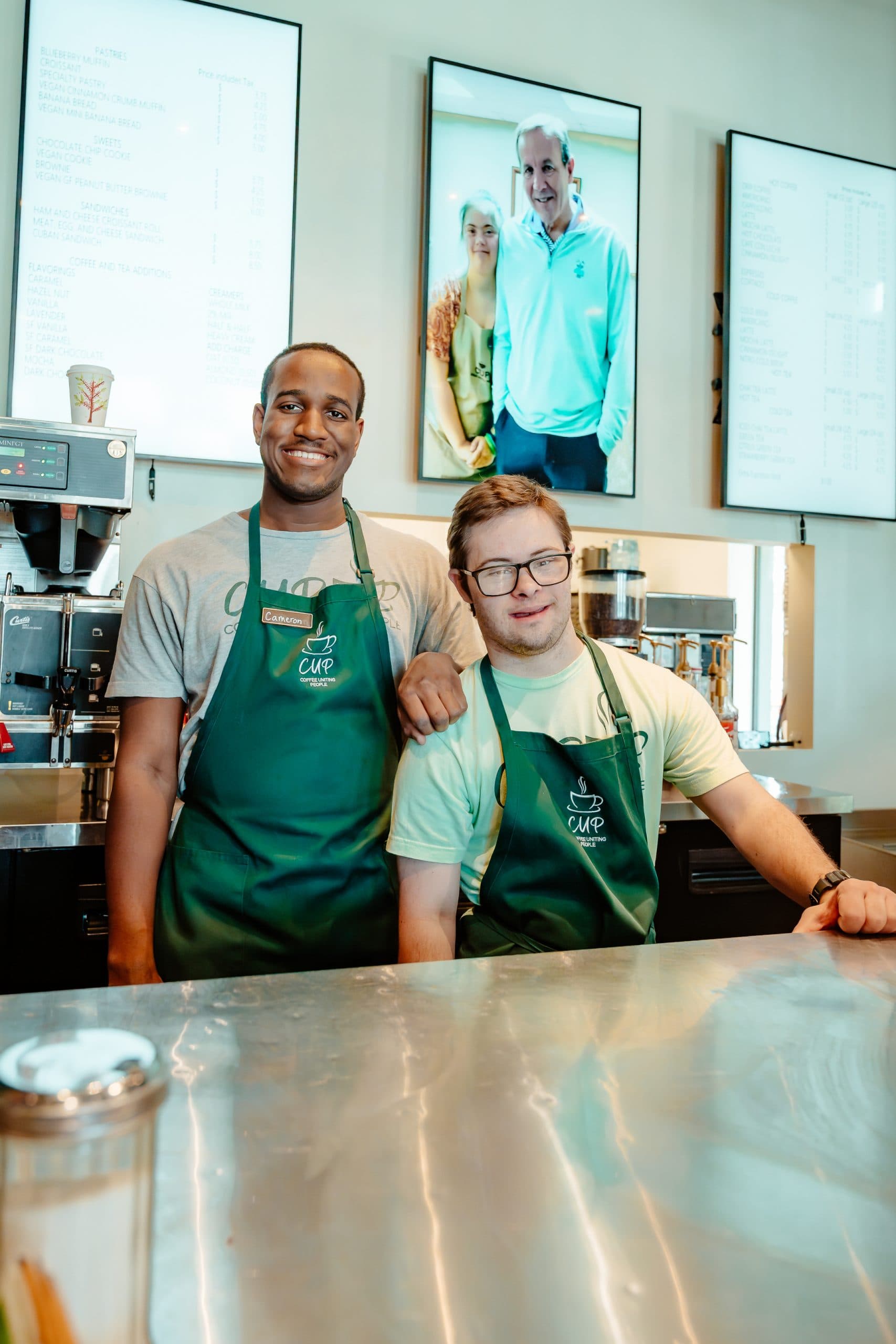 Two CUP baristas in green CUP aprons behind the coffee bar, with a framed portrait of founders Greg and Laura Jones on the wall behind them