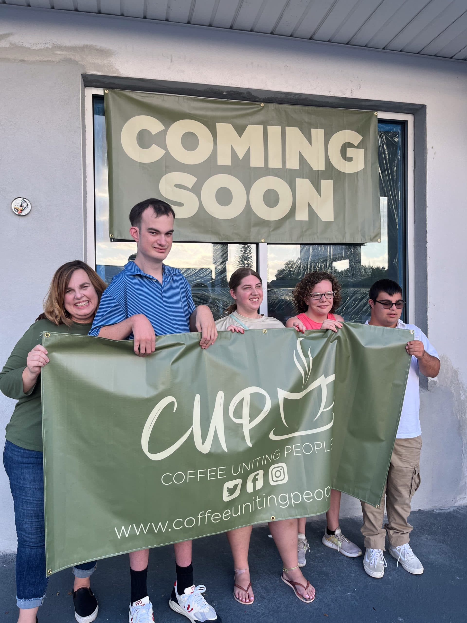 Five CUP team members holding a CUP banner outside the original location, with a 'Coming Soon' banner hanging behind them